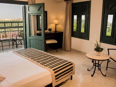 Helles Hotelzimmer mit offenem Tür zu Balkon, Bett und Sitzbereich mit Tischen.