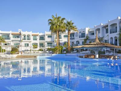 Hotel mit weißer Fassade, mehreren Stockwerken und großem Pool mit Palmen im Vordergrund.