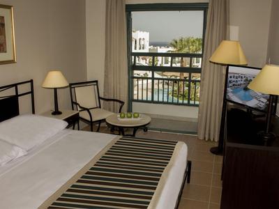 Hell eingerichtetes Hotelzimmer mit Bett, Sitzbereich und Blick auf Pool und Gebäude.