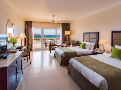 Camera d'hotel luminosa con due letti matrimoniali, TV e balcone con vista mare.