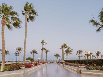Ampio viale lungo una piscina con palme e vista sul mare sotto un cielo limpido.