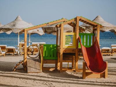 Parco giochi in legno con scivolo sulla spiaggia davanti a lettini e ombrelloni.