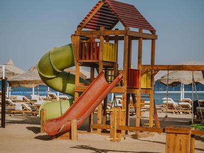 Parco giochi per bambini con scivoli in legno e tunnel verde, circondato da lettini sulla spiaggia.