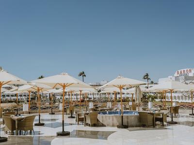 Area esterna dell'hotel con sedute, ombrelloni e palme sotto un cielo sereno.