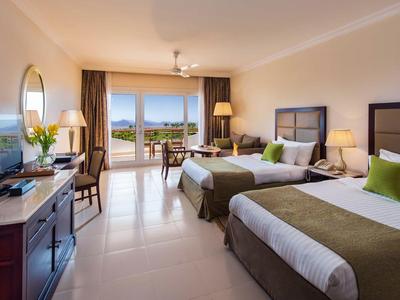 Camera d'hotel luminosa con due letti, TV, scrivania e balcone con vista.