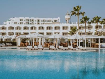 Un grande hotel bianco con molti balconi, palme e una piscina azzurra chiara in primo piano.