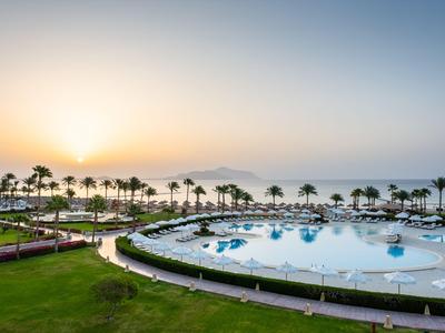 Area piscina di un resort con palme e tramonto sul mare sullo sfondo.
