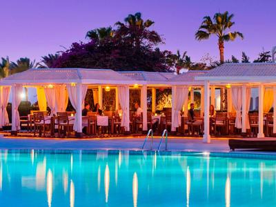 Vista serale di un hotel illuminato con piscina, circondato da palme e cielo viola.