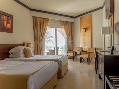 Hotelzimmer mit zwei Einzelbetten, hellem Boden, großem Fenster und warmen Holzmöbeln.