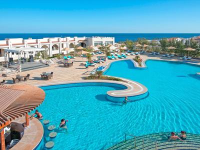 Großer Hotelpool mit Liegestühlen, Sonnenschirmen und Blick auf das Meer im Hintergrund.