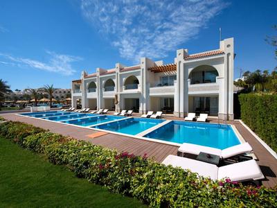 Luxuriöses Hotel mit Pool und Liegestühlen, umgeben von grünen Pflanzen und blauem Himmel.