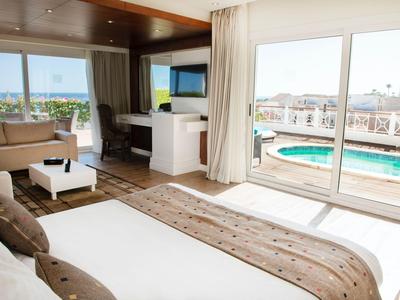 Helles Hotelzimmer mit großem Bett, Sitzecke und Terrasse mit Poolblick.