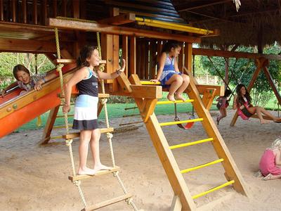 Kinder spielen auf einer hölzernen Kletterstruktur mit Rutsche im Sandbereich eines Hotels.