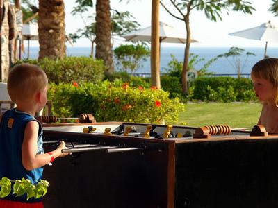 Zwei Kinder spielen an einem Foosball-Tisch im Freien mit Blick auf das Meer und Palmen.