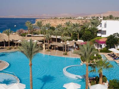 Großer Hotelpool umgeben von Palmen, Sonnenliegen und Blick auf das Meer bei klarem Himmel.