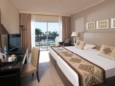 Helles Hotelzimmer mit Doppelbett, Schreibtisch, Fernseher und Balkonblick auf Palmen und Wasser.