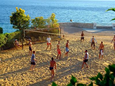 Menschen spielen Volleyball auf einem Sandplatz mit Meerblick bei Sonnenlicht.