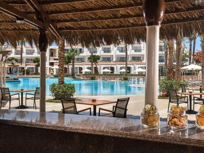 Blick von einer Bar auf einen Pool mit Stühlen und Sonnenschirmen in einer Hotelanlage.