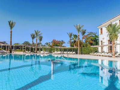 Großer Hotelpool mit klar blauem Wasser, Palmen und Liegestühlen bei sonnigem Himmel.
