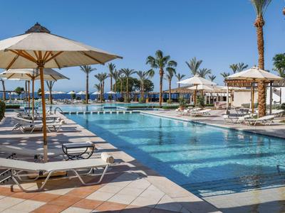 Moderner Poolbereich mit Liegestühlen und Sonnenschirmen in einem Hotelresort unter Palmen.