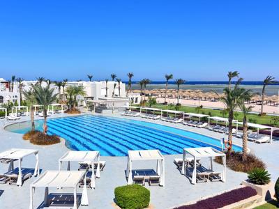 Großer Außenpool mit blauen Liegestühlen und weißen Tischen in einer Hotelanlage am Meer.