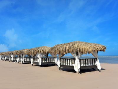 Mehrere überdachte Liegebetten stehen auf einem breiten Sandstrand unter blauem Himmel.