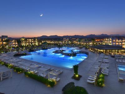 Großer beleuchteter Swimmingpool in einem Hotelresort bei Nacht mit Bergen im Hintergrund.