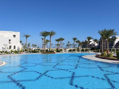 Großer Pool mit blauem Wasser und weißem Hotelgebäude im Hintergrund unter klarem Himmel.