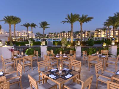 Abendliche Außenansicht eines Resorts mit Holztischen, Stühlen und Palmen am Wasser.