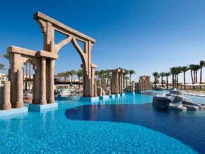 Großer Hotelpool mit blauer Wasserfläche, Palmen und antiken Steinbögen bei klarem Himmel.