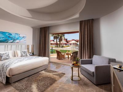 Helles Hotelzimmer mit großem Bett, Sessel, TV und Zugang zu Terrasse mit Gartenblick.