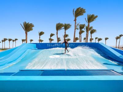 Surfer nutzt Wellen-Simulator im Freien mit Palmen und blauem Himmel im Hintergrund.