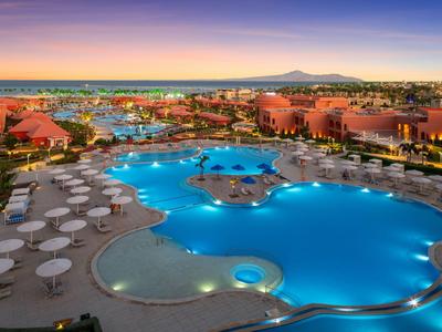 Großer beleuchteter Hotelpool mit Sonnenschirmen, umgeben von roten Gebäuden bei Sonnenuntergang.