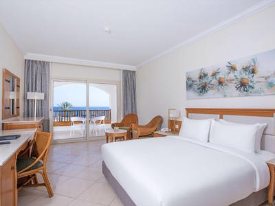 Helles Hotelzimmer mit Doppelbett, Schreibtisch, Sesseln und Balkon mit Meerblick.