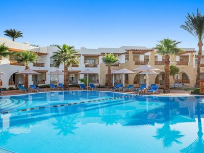 Großer Pool vor zweistöckigen Hotelgebäuden und Palmen unter klarem blauem Himmel.