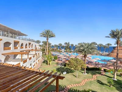 Hotelanlage mit Garten, Palm Bäumen, Pool und Sonnenschirmen unter klarem blauem Himmel.