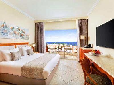 Helles Hotelzimmer mit großem Bett und Balkon mit Meerblick bei klarem Himmel.