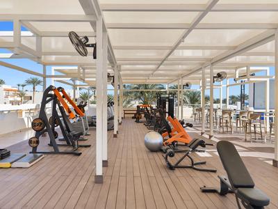 Freiluft-Fitnessbereich mit modernen Geräten unter weißem Sonnendach und Blick auf Palmen.