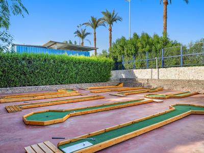 Minigolfplatz mit grünen Bahnen, roten Wegen, Holzbänken und Palmen im Hintergrund.