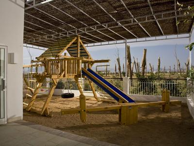 Überdachter Spielplatz mit Rutsche, Schaukeln und Sandfläche neben Hotelgebäuden.