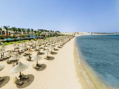 Breiter Sandstrand mit zahlreichen Strohmsonnenschirmen und Palmen am Hotelgelände unter blauem Himmel.