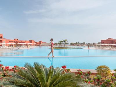 Eine Frau geht am Rand eines großen Pools in einer Resortanlage mit roten Gebäuden und Palmen.