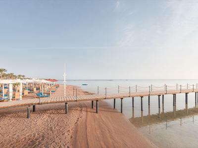Sandstrand mit Liegestühlen und einem langen Steg über ruhigem Wasser bei klarem Himmel.