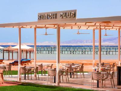 Offener Strandclub mit Holzdach und Tischen mit Blick auf das Meer und eine lange Pier.
