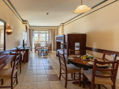 Camera d'hotel luminosa con zona pranzo, sedie in legno, tavolo, pavimento piastrellato e accesso al balcone.