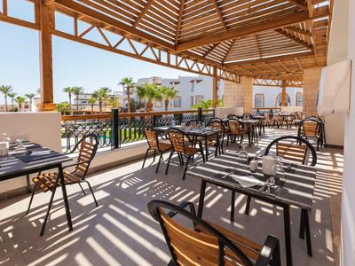 Area ristorante aperta dell'hotel con tavoli, sedie e vista sulla piscina e le palme.
