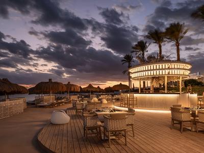 Terrasse d'hôtel au crépuscule avec tables, chaises et bar éclairé sous un ciel nuageux.