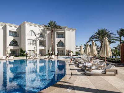 Hotel con grande piscina, lettini e ombrelloni davanti a edificio chiaro e cielo blu.