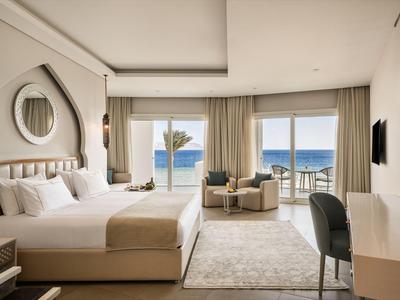 Chambre d'hôtel luxueuse avec lit double, coin salon et vue sur la mer par de grandes fenêtres.