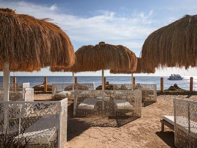 Spiaggia con ombrelloni di paglia e sedute bianche di fronte al mare sotto cielo azzurro.